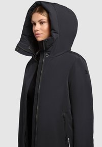 Veste isolante noire avec une grande capuche, fermeture éclair à l'avant et poches latérales. Texture lisse avec un design minimaliste et sans motifs visibles.