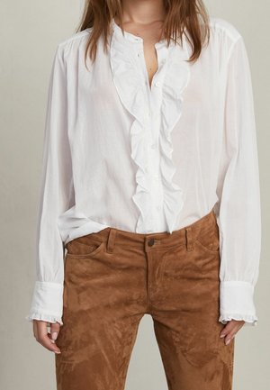 Femme portant un chemisier blanc à manches longues avec des volants, rentré dans un pantalon en daim marron taille haute, debout, les mains détendues.