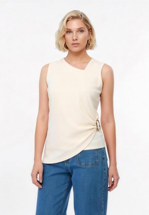 Mujer rubia con top sin mangas color crema con detalle asimétrico de hebilla en el lateral y jeans azules, contra fondo blanco.