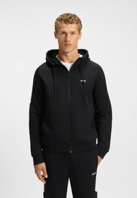 Czarna bluza z kapturem zapinana na zamek, wykonana z gładkiego materiału, z kapturem, sznurkami do regulacji i minimalistycznym logo na klatce piersiowej. Ściągacze przy mankietach i u dołu.