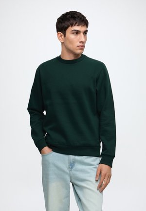 Jeune homme portant un sweat-shirt vert foncé à col rond et un jean bleu clair, debout avec une main dans la poche, regardant sur le côté.