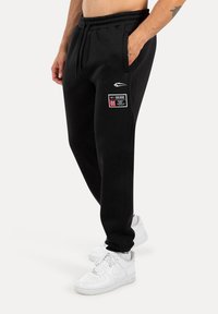 Schwarze Jogginghose aus Baumwollmischung mit elastischem Bund, seitlichen Taschen und Logo-Detail. Kombiniert mit weißen Turnschuhen.