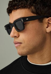 Lunettes de soleil rectangulaires noires avec des montures épaisses, présentant des accents en argent sur les branches, portées par une personne avec des cheveux courts et bouclés.