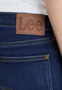 Jean en denim bleu foncé avec une patch en cuir brun affichant "Lee" en relief. Présente des coutures orange le long des coutures et des poches.