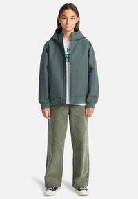 Giacca impermeabile verde con cappuccio, cerniera frontale e tasche laterali, indossata sopra una t-shirt grigia e pantaloni larghi verde oliva.
