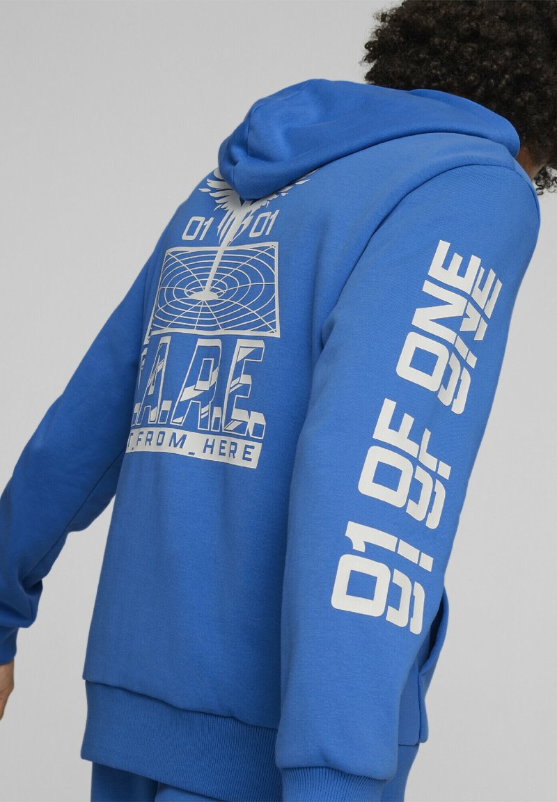 Personne portant un sweat à capuche bleu avec un texte graphique blanc "01 OF ONE" sur la manche et "A.P.E. FROM_HERE" dans le dos sous un motif géométrique.