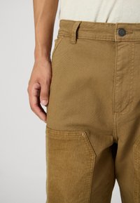 Pantalons en velours côtelé brun avec une finition en tissu lisse, poches latérales assorties et une fermeture à bouton à la taille, agrémentés de détails de couture subtils.