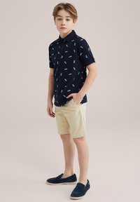 Tengerészkék póló, amit világoskék minták színesítenek, bézs rövidnadrággal párosítva. A fiú fekete slip-on cipőt visel.