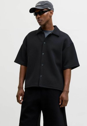 Homme portant une chemise noire à manches courtes boutonnée, un short noir, une casquette noire et des lunettes de soleil sur un fond clair uni.