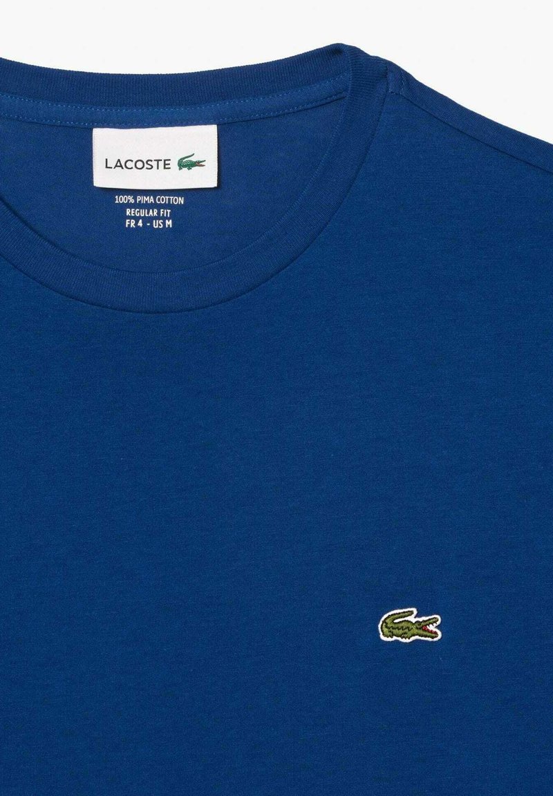 Blaues T-Shirt aus 100% Pima-Baumwolle, mit Rundhalsausschnitt und einem kleinen grünen Lacoste-Logo, das auf der linken Brust gestickt ist. Normale Passform.
