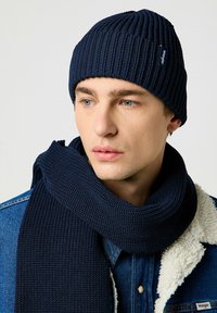 Navy blue knit beanie and scarf set, featuring ribbed texture, worn over a denim jacket with a shearling collar. Subtle logo on the beanie.
