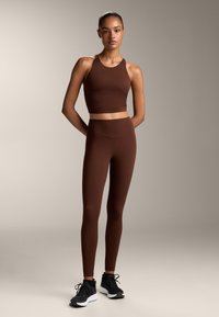 Hnědý crop top a legíny s vysokým pasem vyrobené z elastického materiálu, doplněné černými sportovními botami. Jednoduchý design bez vzorů nebo akcentů.