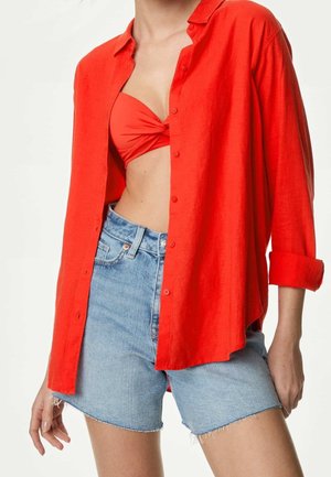 Kobieta nosząca rozpiętą czerwoną koszulę na czerwonym bandeau z przednim skrętem i jasnoniebieskie dżinsowe krótkie spodenki z postrzępionymi krawędziami.