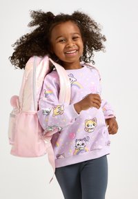 Lila Sweatshirt mit Cartoon-Tiermustern, graue Leggings und ein glänzender pinker Rucksack mit einer Pom-Pom-Akzent. Kind lächelt und posiert.