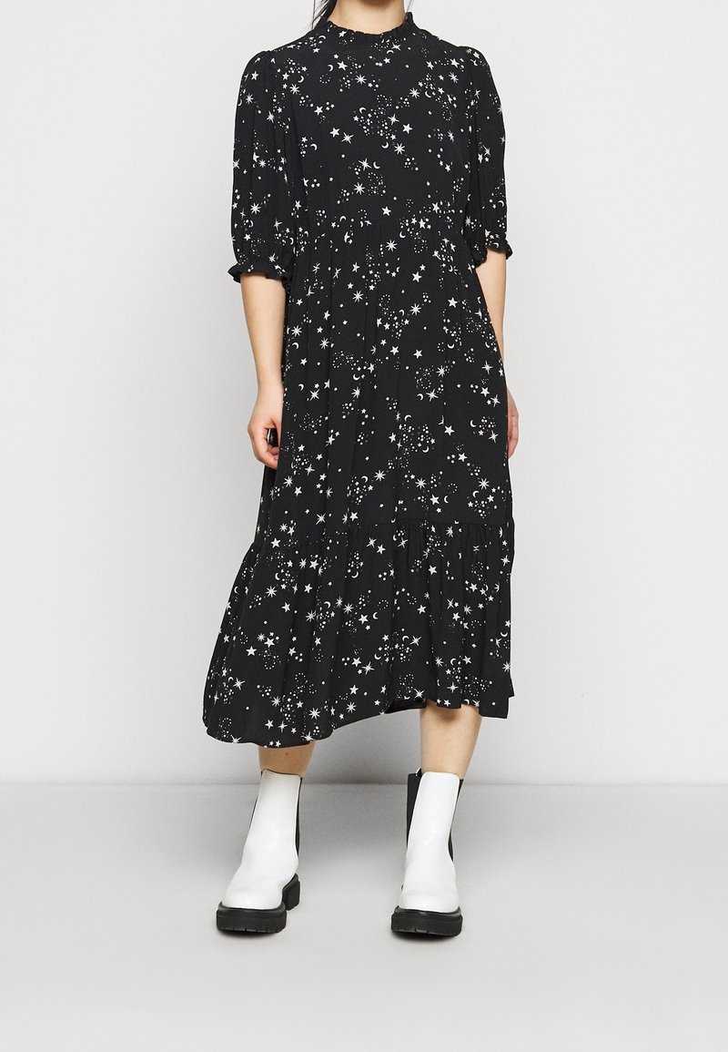 Femme portant une robe midi noire avec des motifs d’étoiles et de lunes blancs et des bottines blanches avec des semelles noires, debout devant un fond uni.