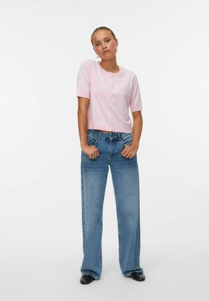 Vrouw in een lichtroze kortebloesje met knopen, losse blauwe jeans en zwarte schoenen, staand met handen in de zakken op een witte achtergrond.