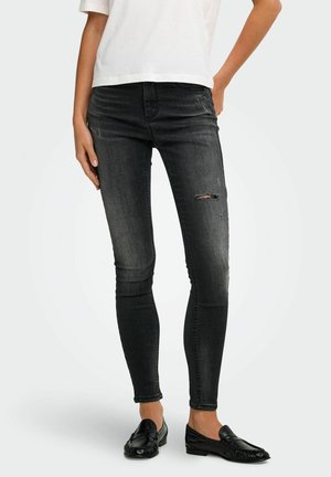 Femme portant un jean skinny noir déchiré, une chemise blanche et des mocassins noirs, debout avec une main sur la hanche et l'autre relâchée.