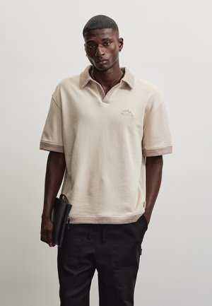 Homme portant un polo beige à manches courtes avec col et poignets texturés, pantalon noir, tenant une pochette noire zippée sur un fond uni.