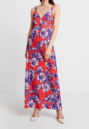 Seorang wanita mengenakan gaun maxi tanpa lengan berwarna merah dengan pola bunga ungu dan pink, serta sepatu hak tinggi putih di latar belakang polos.