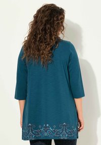 Teal tunika lavet af blødt stof, med 3/4 ærmer og indviklet blå blomsterbroderi i kanten. Glat tekstur, afslappet design.