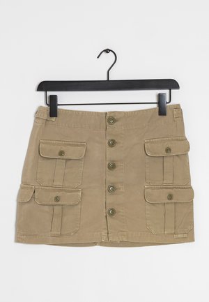Beige mini-rok met knopen aan de voorkant en vier klepzakken, hangend aan een zwarte hanger tegen een witte achtergrond.