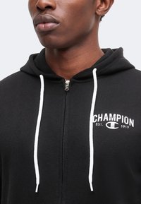Κοντινό πλάνο ατόμου που φοράει μαύρο Champion hoodie με λευκά κορδόνια και λογότυπο στο στήθος, το φερμουάρ είναι μερικώς κλειστό.