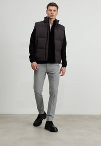 Black puffer vest with a high collar, worn over a black sweater, paired with light gray trousers and black sneakers. Smooth texture, minimal design.
