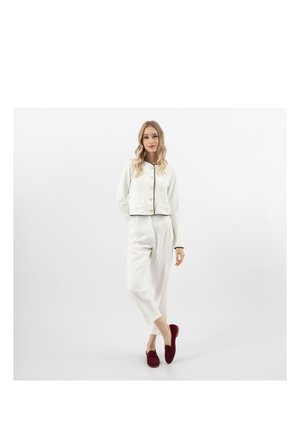 Vrouw staat met gekruiste benen, draagt een witte jas met knopen en een broek, gecombineerd met donkerrode instapschoenen, tegen een effen witte achtergrond.