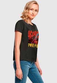 Black cotton t-shirt featuring red and yellow AC/DC graphic with "PWR" in lightning style. Short sleeves, relaxed fit, crew neck design.