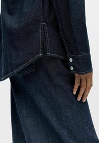 Donkerblauw denim overhemd met een losse pasvorm. Voorzien van witte stiksels, zijsplitten en twee knopen aan de manchetten voor verstelbaarheid.