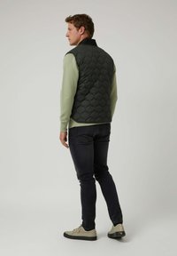 Chaleco acolchado de color verde oscuro sin mangas, combinado con una camisa de manga larga verde claro, jeans negros y zapatillas beige. Vista trasera, fondo liso.