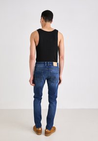 Jeans de denim azul con un acabado текстurizado, que presentan un parche de cuero marrón en la parte trasera, bolsillos traseros con detalles de costura, combinados con una camiseta sin mangas negra.