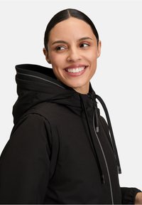 Femme aux cheveux foncés dans une veste à capuche noire sourit en regardant à droite sur un fond gris uni.