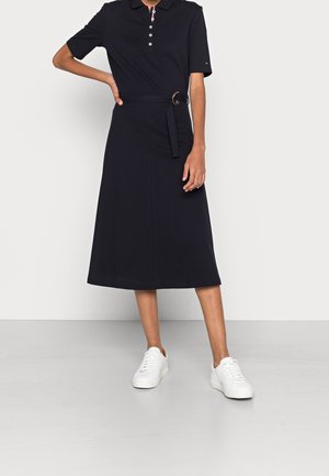 Personne portant une robe noire à manches courtes avec une ceinture et des baskets blanches, debout, une main sur la hanche, devant un fond uni.