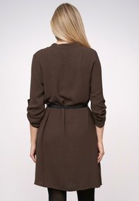 Braunes, knielanges Kleid aus strukturiertem Stoff, mit tailliertem Gürtel und Dreiviertelärmeln. Rückansicht ohne Muster oder Verzierungen.