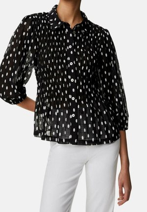 Zwarte doorzichtige blouse met witte ovale polkadots, knoopsluiting aan de voorkant, driekwartmouwen met pofeffect, gedragen met witte broek.