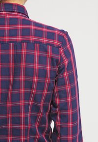 Chemise à carreaux bleue et rouge avec un tissu texturé. Elle a un col boutonné et des détails de plis au dos.