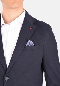 Blazer blu navy con texture, spilla a forma di forbice rossa, camicia bianca e fazzoletto da taschino blu a fantasia. Chiuso con il risvolto visibile e design elegante.