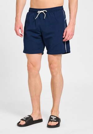 Mand iført marineblå badeshorts med hvid snor og sorte Hummel sliders mod en ensfarvet baggrund.