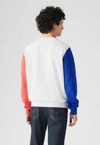 Man met krullend haar in een wit sweatshirt met een rode linkermouw en een blauwe rechtermouw, gecombineerd met donkerdenim jeans, staande in profiel.