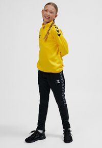 Gul atletisk pullover med hög krage och svarta chevron-accenter. Svarta joggerbyxor med grå sidolinjer och svarta atletiska skor.