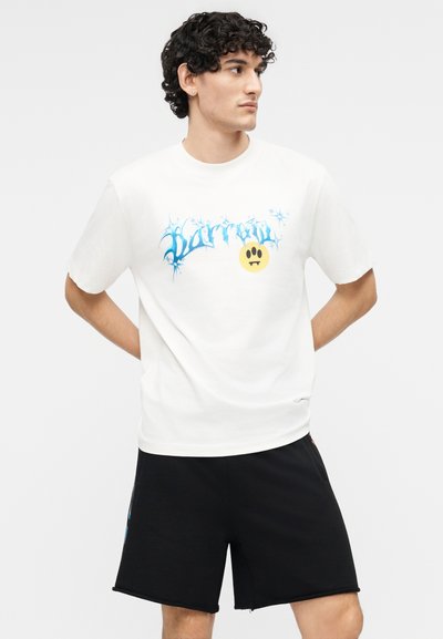 Νεαρός με μαλλιά σγουρά που φοράει λευκό μπλουζάκι με μπλε κείμενο και κίτρινο emoji τύπου βαμπίρ, σε συνδυασμό με μαύρο σορτς, κοιτάζει δεξιά.