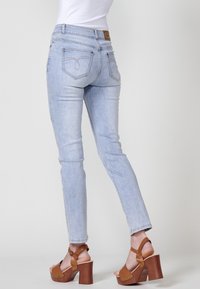Personne portant un jean skinny bleu clair et des sandales à talons hauts marron, vue de dos sur un fond gris uni.