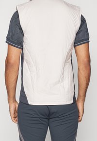 Chaleco sin mangas de color beige claro, acolchado con un acabado suave, presenta un escote redondeado y un corte ajustado, usado sobre una camisa oscura con estampado.