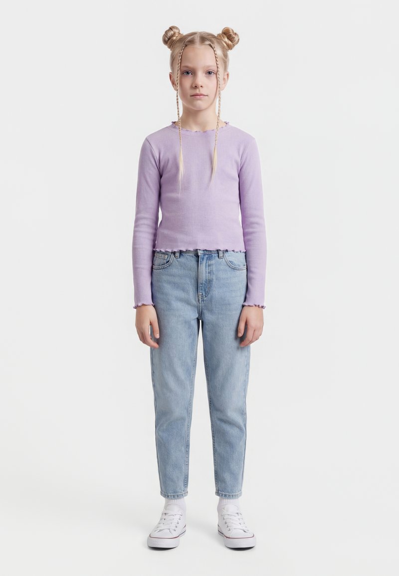 Mergaitė su blondinėmis dviem kuodeliais ir kasomis vilki levandų spalvos ilgomis rankovėmis marškinėlį, šviesiai mėlynus džinsus ir baltus sportinius batelius, stovinti veidu į priekį.