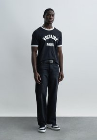 Νεαρός άνδρας στέκεται μπροστά σε απλό λευκό τοίχο φορώντας μαύρο μπλουζάκι "Voltaire Paris", μαύρο τζιν, μαύρα και λευκά αθλητικά παπούτσια και κολιέ με χάντρες.