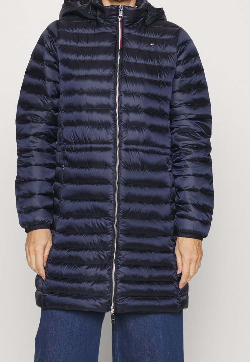 Tummansininen tikattu toppatakki huppulla, edessä vetoketjukiinnitys ja joustavat hihansuut. Valmistettu kiiltävästä, kevyestä materiaalista.