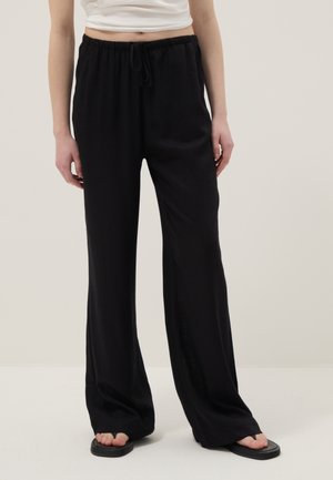 Persoon die losse zwarte broek met trekkoord, een kort wit topje en zwarte sandalen met vierkante neus draagt, staand tegen een effen achtergrond.