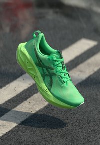 Grüner Sportschuh mit einem strukturierten Mesh-Obermaterial, leichtem Sohlenprofil, schwarzen Akzenten und runder Zehenpartie, in der Luft über einer Asphaltoberfläche dargestellt.