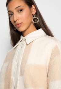 Beige en crème geblokt fleece jack met een knoopsluiting op de kraag en een zachte textuur; model draagt grote ronde geprofileerde oorbellen.